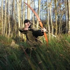 Jan van der Veen schiet een zelfgemaakte houten pijl en boog vanuit het hoge gras, gebaseerd op het boek Houtkoorts: Zelf een handboog maken.