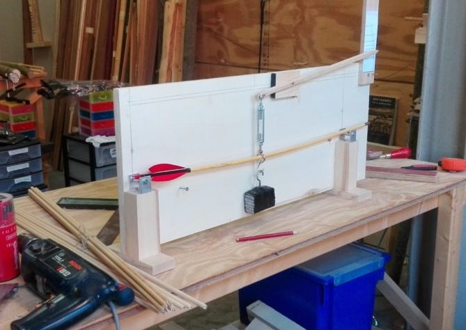 Een pijl wordt met gewichten getest op buiging. De pijl is zelf van hout te maken, met veren en een nok.
